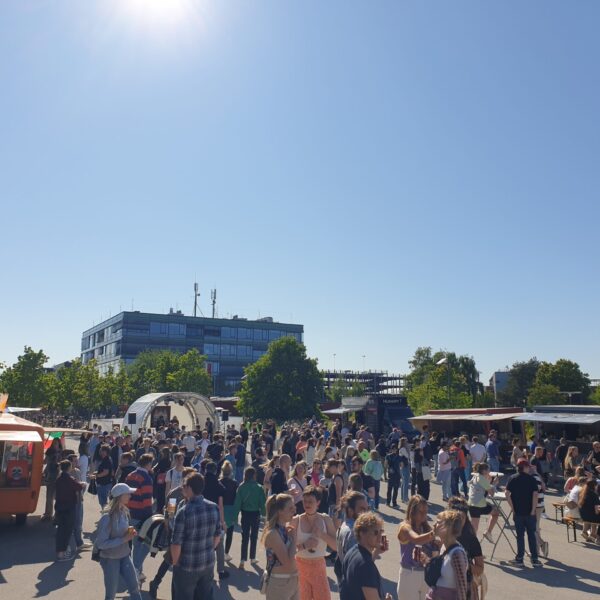 Es sind verschiedene Essensstände zu sehen vor denen Menschen stehen oder an Biertischen sitzen.