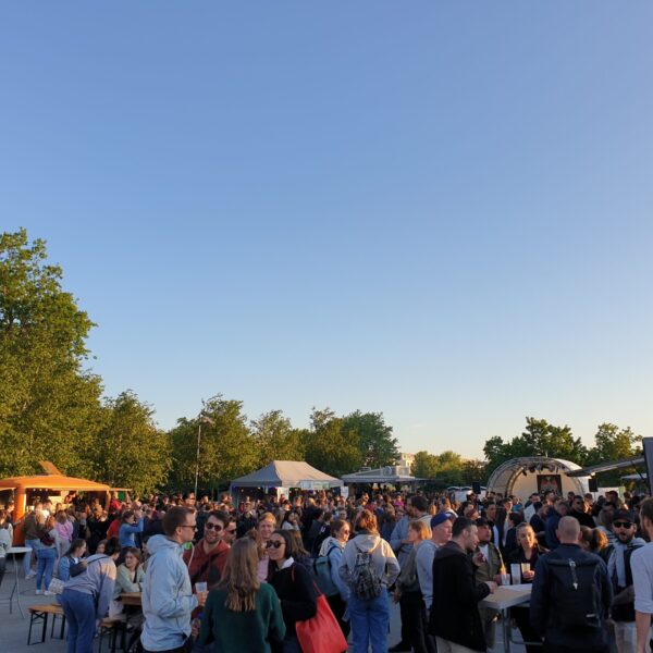 Menschen die an Biertischen sitzen oder davor in Gruppen stehen. Der Himmel ist strahlen blau.