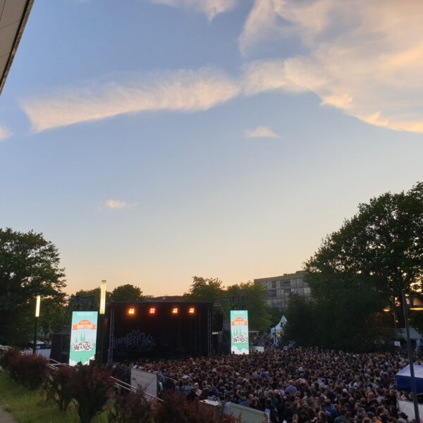 Bild von einem Hügel aus das den Sonnenuntergang zeigt. am unteren Rand sieht man einen Menschenmenge die vor einer Bühne steht.