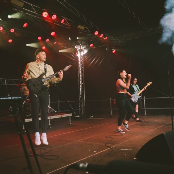 Drei Musiker*innen auf einer beleuchteten Bühne beim Singen und Gitarrespielen.