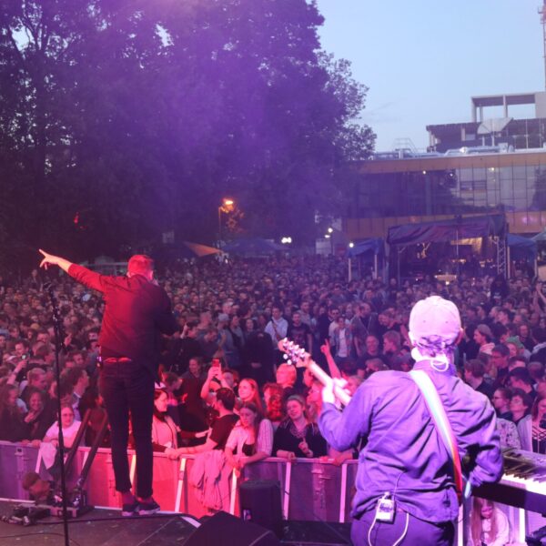 Menschenmenge von der Bühne aus Fotografiert. 2 Personen stehen im Vordergrund auf der Bühne. Die rechte spielt Gitarre und die linke singt.
