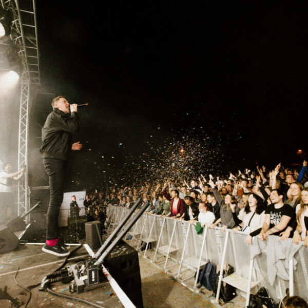 Bild aus dem Breich vor der Bühne von der ersten Reihe die vor der Bühne steht. eine Person steht auf der Bühne und singt