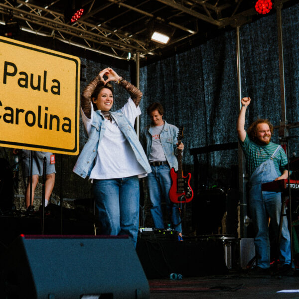 Auf der Bühne stehen 3 Personen und auf einem Ortseingangsschild steht Paula Carolina.